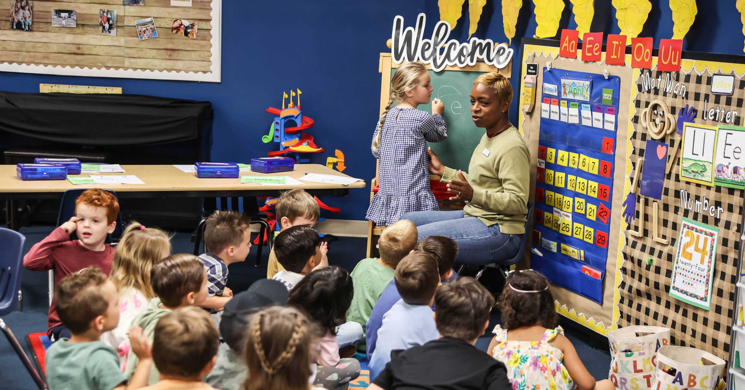 Teacher helping a young student at the classroom board under a Welcome sign