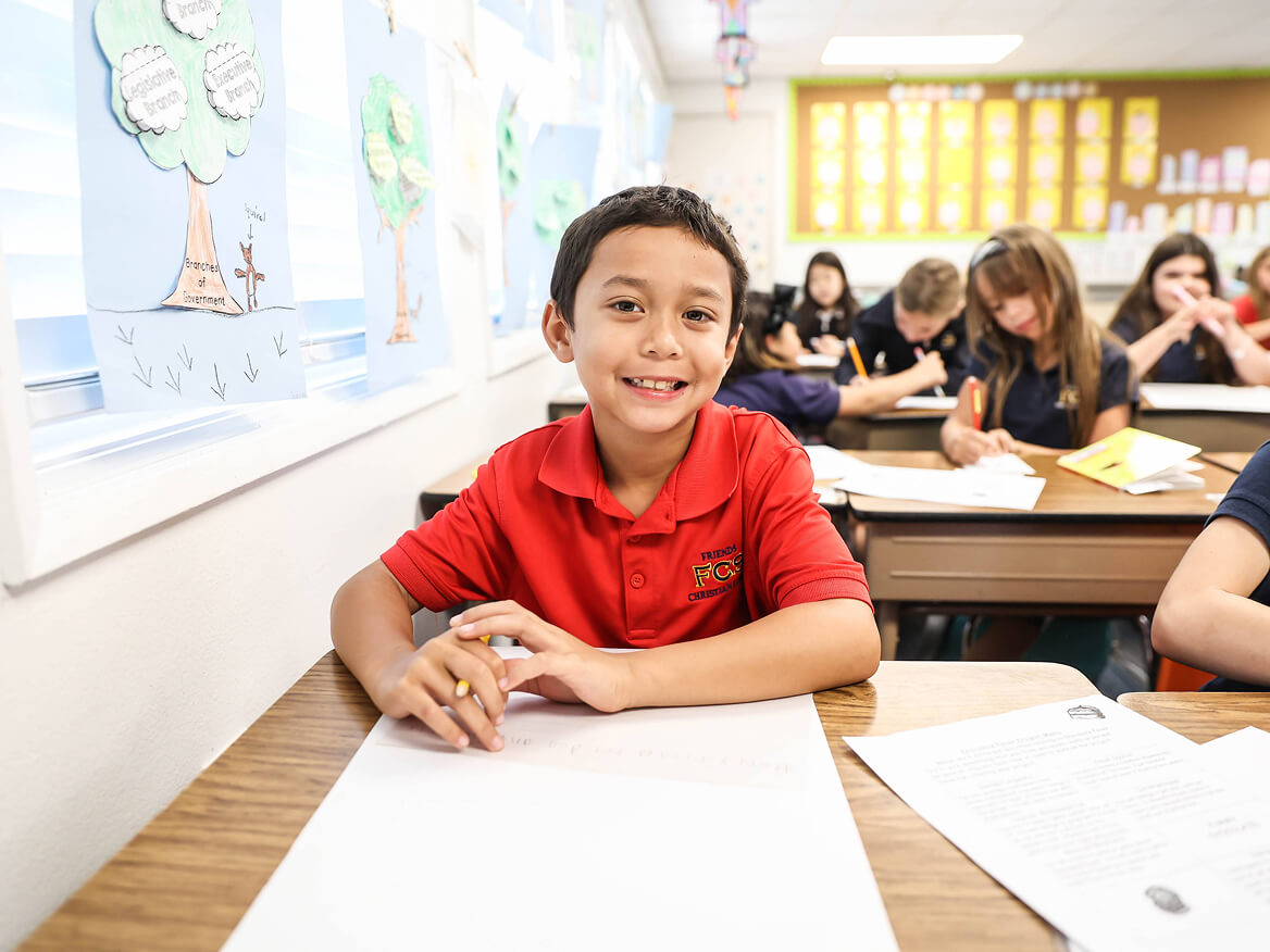 Smiling boy in a red uniform shirt sitting at his classroom desk