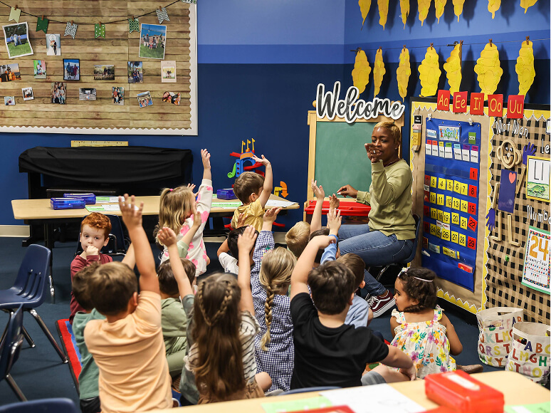 Students sitting on the classroom rug raising their hands during a lesson