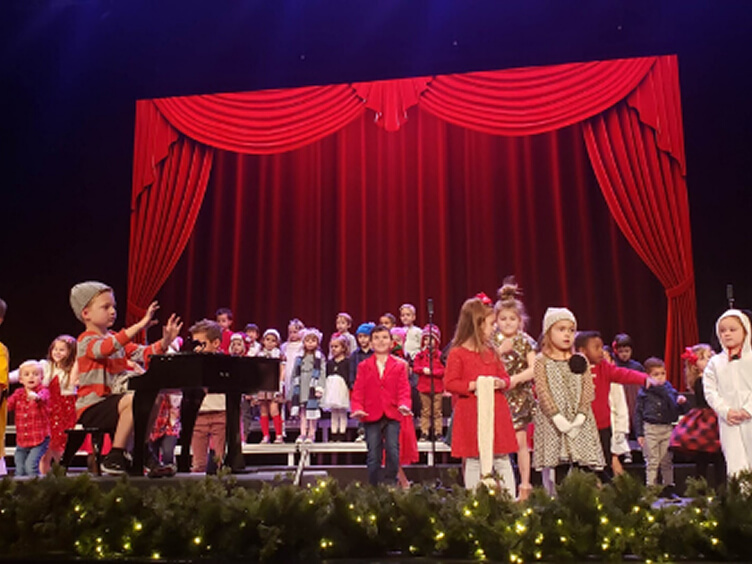 Young students in holiday costumes performing at a Christmas program in front of a red curtain
