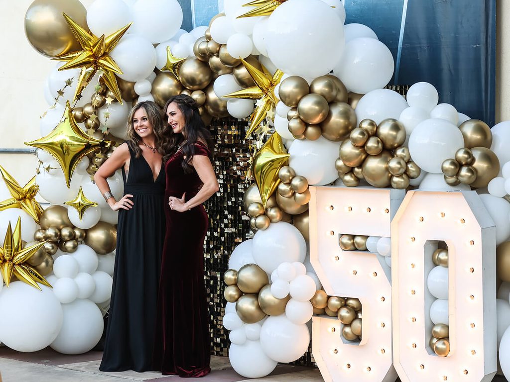 Two women posing in front of a gold and white balloon display for the 50th anniversary