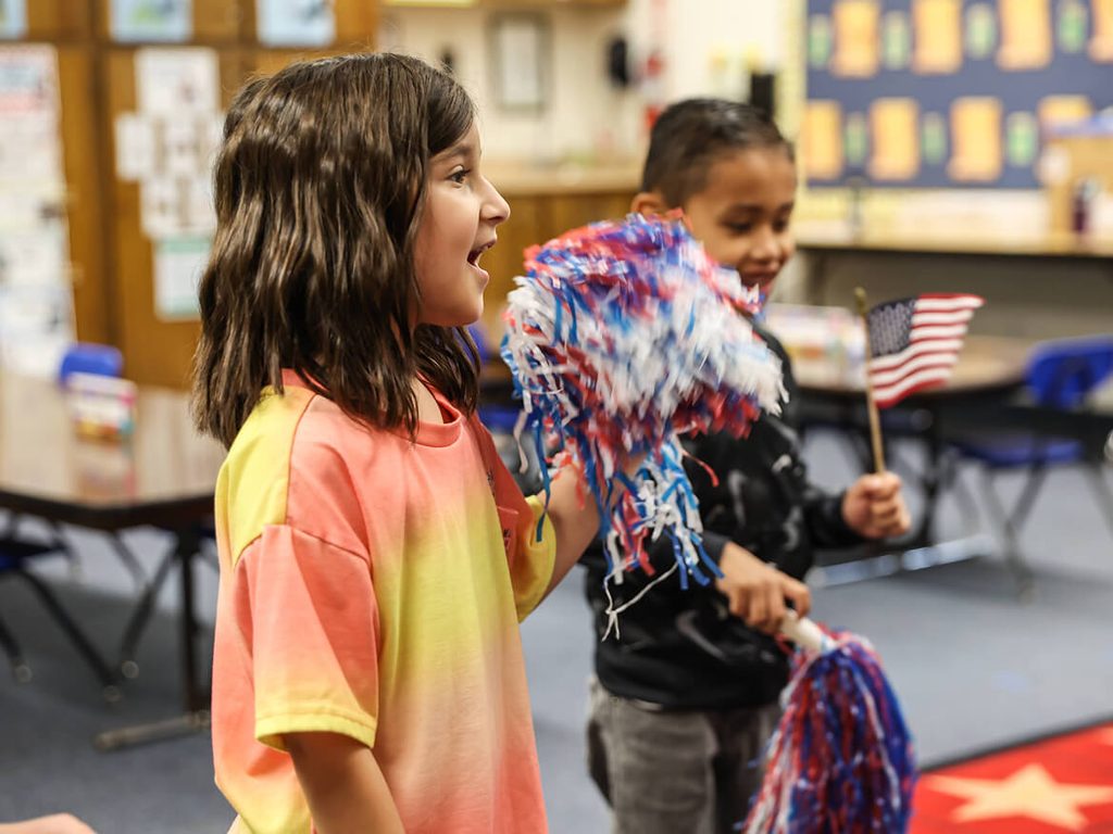 Smiling students waving red, white and blue pom-poms in the classroom