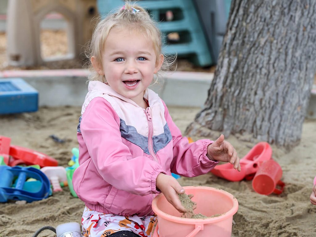Smiling toddler playing with sand toys in the preschool sandbox